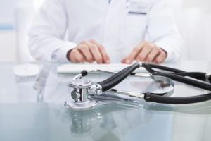 Conceptual medical and healthcare image with a close up of a stethoscope lying on a doctors desk with the hands of the doctor visible behind working on a computer